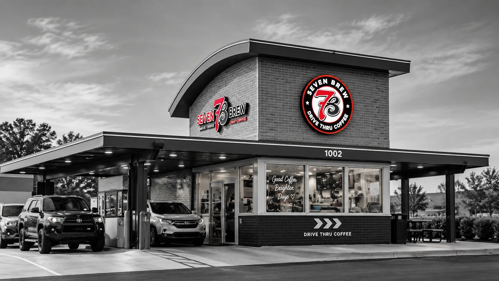 A 7 Brew drive-thru stand with two cars waiting under a lit canopy, the red logo the only color in an otherwise black-and-white scene.