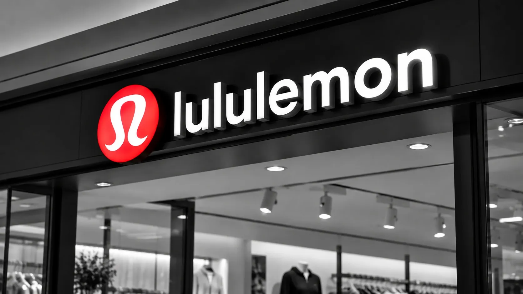 A lululemon storefront sign lit above a darkened shop floor, the red logo the only color in an otherwise black-and-white scene.