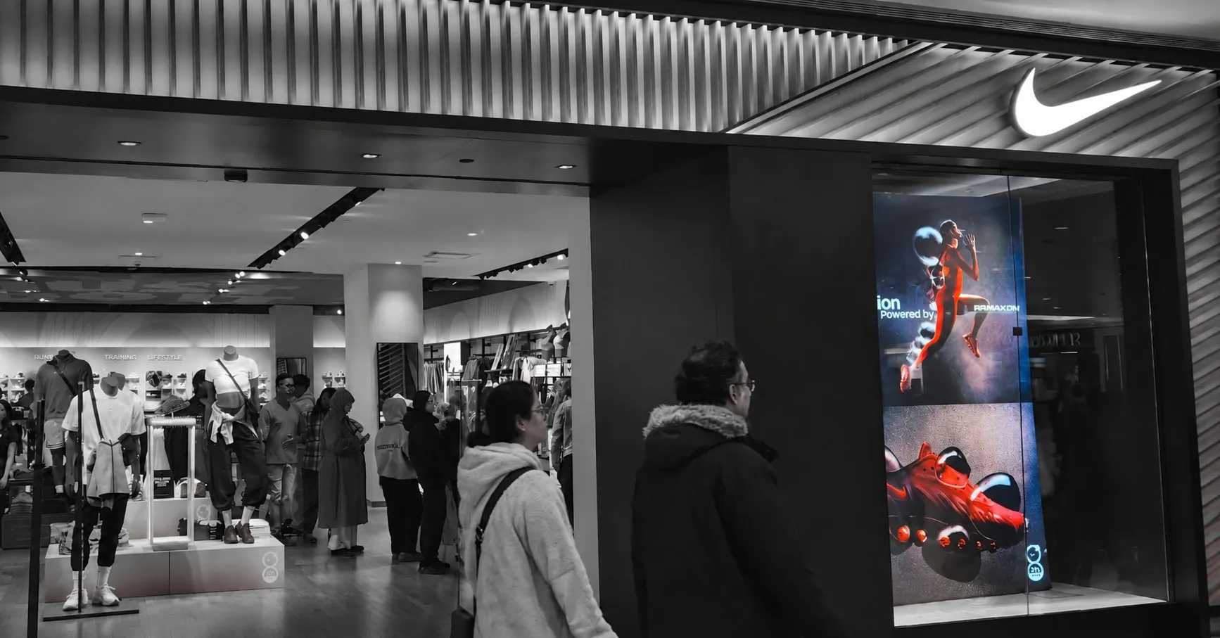A Nike store in a shopping mall, black and white except for a lit advertising screen glowing red beside the entrance. Shoppers browse inside, under wall signs reading Running, Training and Lifestyle.